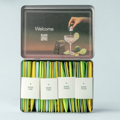 Tin box with green packets labeled 'Week One' to 'Week Four' on a light gray background.